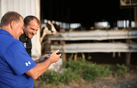 Two men, outside, next to a barn, looking at a tablet